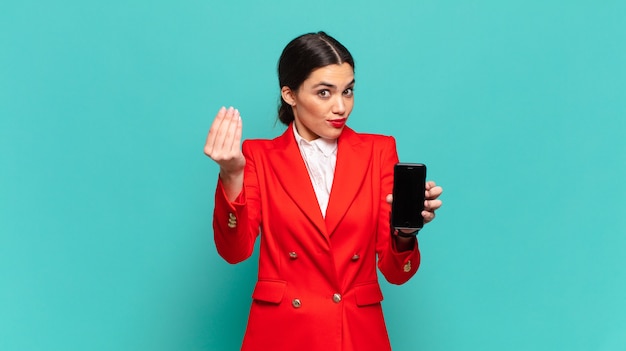 Mulher jovem e bonita fazendo capice ou gesto de dinheiro, dizendo para você pagar suas dívidas !. conceito de telefone inteligente