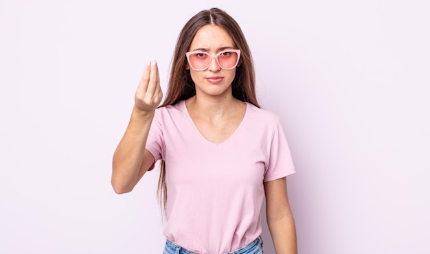 Mulher jovem e bonita fazendo gesto de capice ou dinheiro, dizendo para você pagar. conceito de óculos de sol rosa