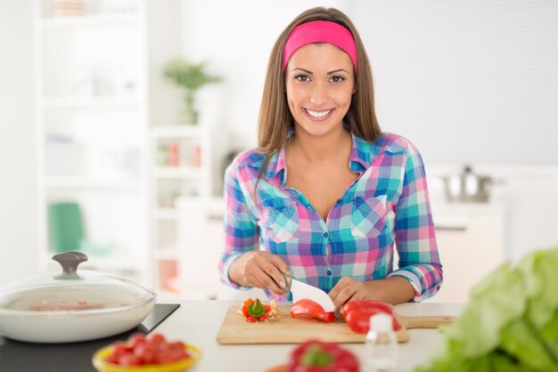 Foto mulher jovem e bonita fazendo refeição saudável na cozinha doméstica. ela está cortando pimenta vermelha na tábua da cozinha. olhando para a câmera.