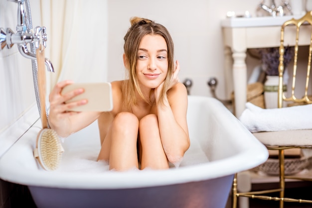 Mulher jovem e bonita fazendo selfie com o telefone sentada na banheira retrô dentro de casa