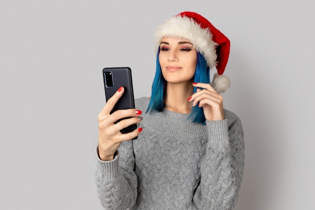 Mulher jovem e bonita feliz com chapéu de papai noel tome selfie sobre fundo cinza. conceito de celebração de ano novo de natal