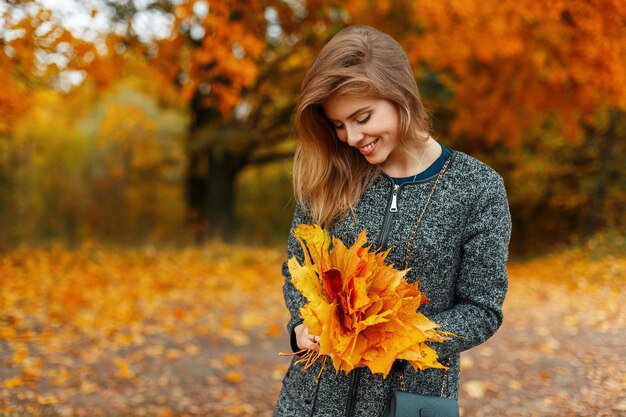 Mulher jovem e bonita feliz com roupas da moda de outono segurando folhas amarelas ao ar livre
