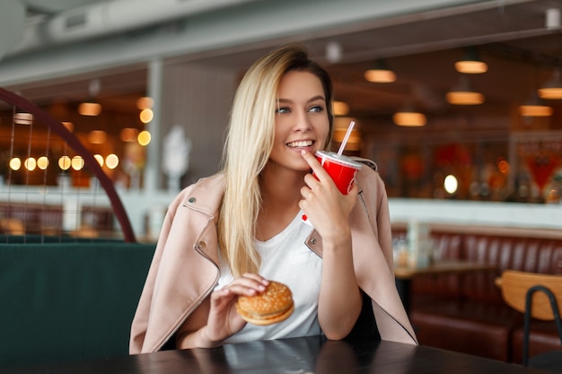 Mulher jovem e bonita feliz com um hambúrguer e uma bebida comendo fast food