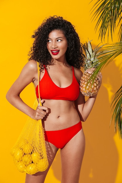 mulher jovem e bonita feliz em trajes de banho posando isolado sobre a parede de parede amarela perto de planta tropical de natureza verde segurando limões e abacaxi.