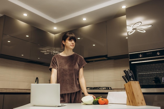 Mulher jovem e bonita feliz gostando de cozinhar procurando uma receita no laptop na cozinha