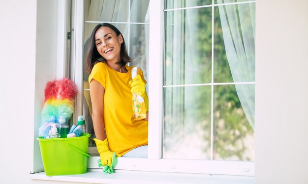 Mulher jovem e bonita feliz usando um espanador e um spray enquanto limpa uma janela da casa