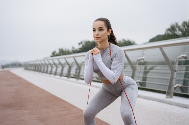 Mulher jovem e bonita fitness em roupas esportivas elegantes treinando ao ar livre, fazendo exercícios de agachamento com banda de resistência