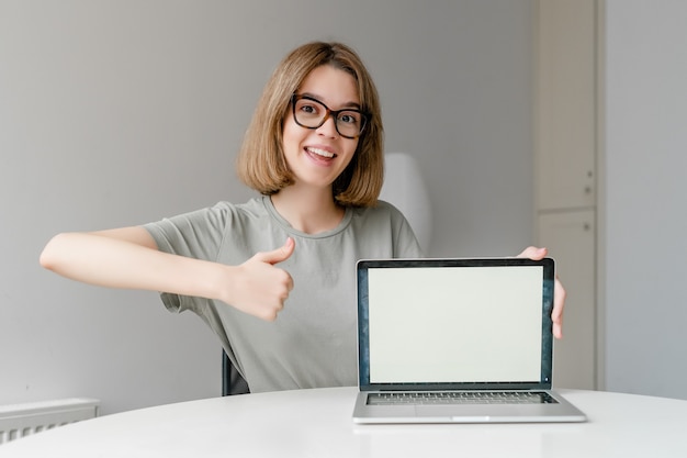 Mulher jovem e bonita Freelancer mostrando a tela de maquete do computador portátil e o polegar para cima sentado em casa. Trabalho autônomo, conceito de educação online