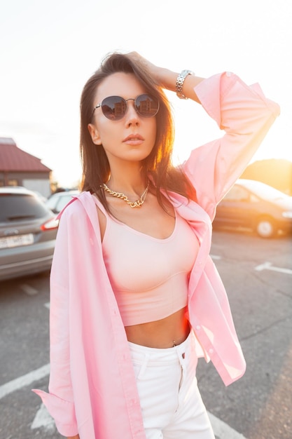 Mulher jovem e bonita glamour com óculos de sol legais em roupas da moda com camisa rosa e regata anda na rua ao pôr do sol Roupa de estilo feminino de verão