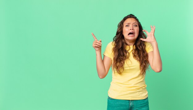 Mulher jovem e bonita gritando com as mãos para o alto e apontando para o espaço da cópia