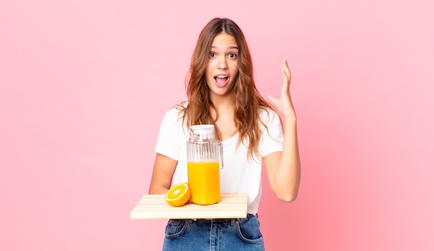 Mulher jovem e bonita gritando com as mãos para o alto e segurando uma bandeja com suco de laranja