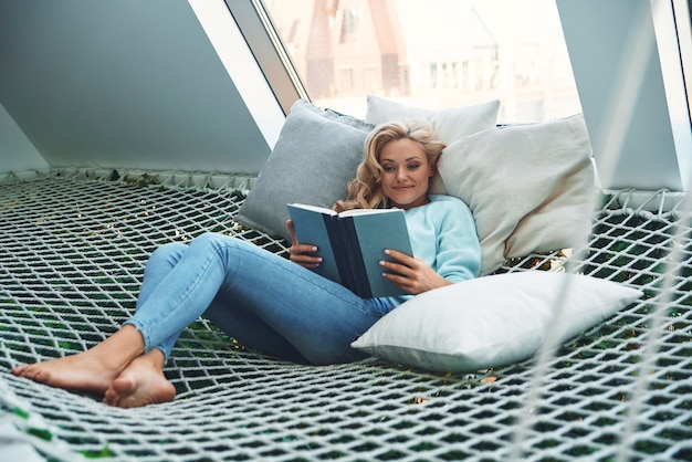 Mulher jovem e bonita lendo livro e sorrindo enquanto relaxa em uma grande rede em casa