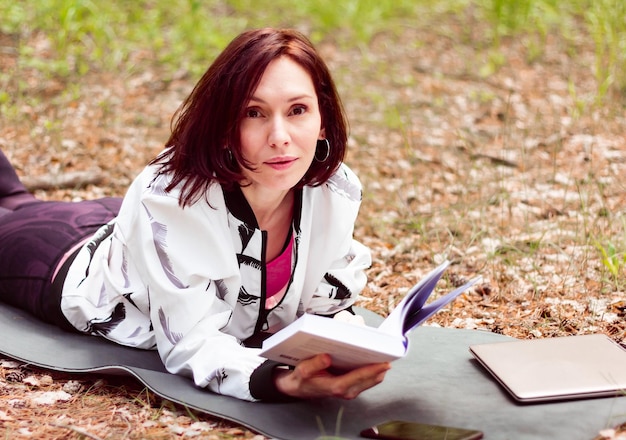 Mulher jovem e bonita lendo um livro no parque enquanto estava deitada de madrugada no tapete de ioga com laptop nas proximidades