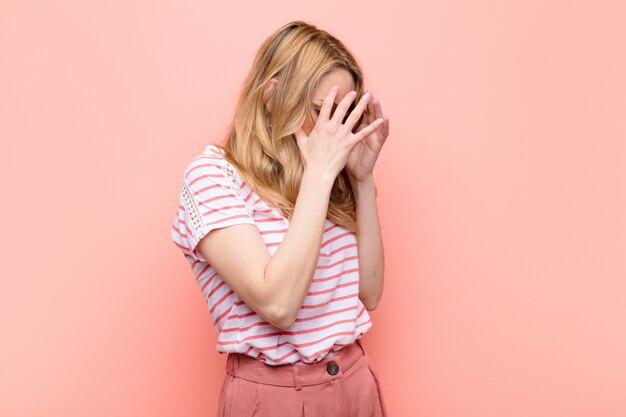 Mulher jovem e bonita loira cobrindo os olhos com as mãos com uma expressão triste e frustrada de desespero, chorando, vista lateral contra uma parede de cor lisa