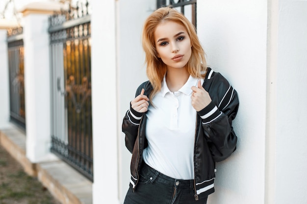 Mulher jovem e bonita loira com uma jaqueta preta e uma camisa pólo branca perto de uma parede branca