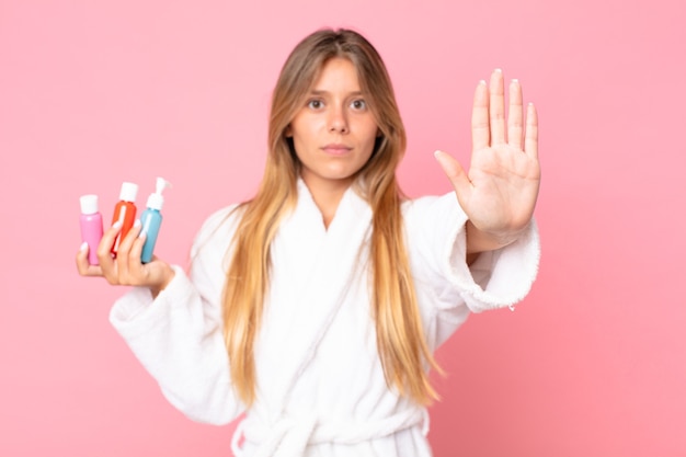 Mulher jovem e bonita loira vestindo roupão de banho e segurando um produto cosmético