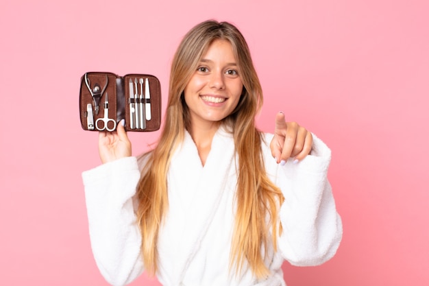 Mulher jovem e bonita loira vestindo roupão de banho e segurando uma bolsa de maquiagem com ferramentas de unhas