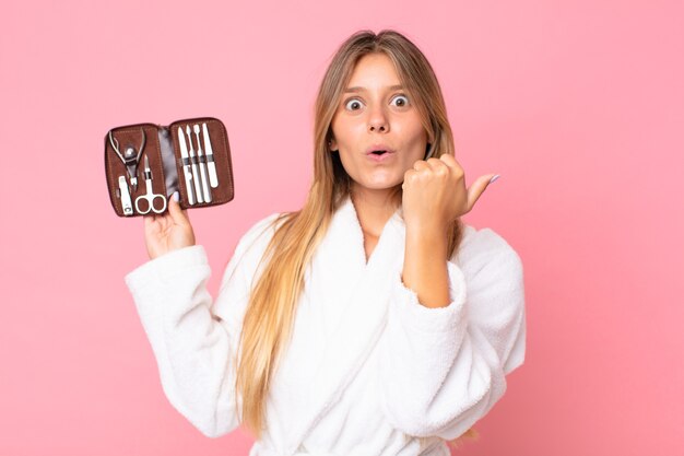 Mulher jovem e bonita loira vestindo roupão de banho e segurando uma bolsa de maquiagem com ferramentas de unhas