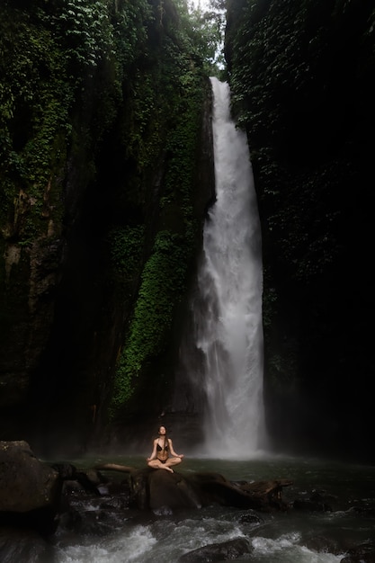 Mulher jovem e bonita meditando na posição de lótus enquanto fazia ioga em uma floresta maravilhosa perto de cachoeira. linda fêmea praticando yoga na rocha perto de cachoeira tropical