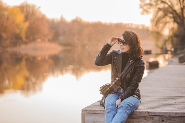 Mulher jovem e bonita moda sentada no cais no pôr do sol com bela vista do rio