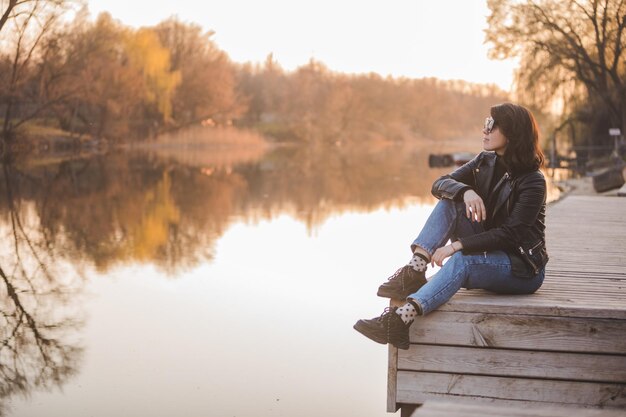 Mulher jovem e bonita moda sentada no cais no pôr do sol com bela vista do rio