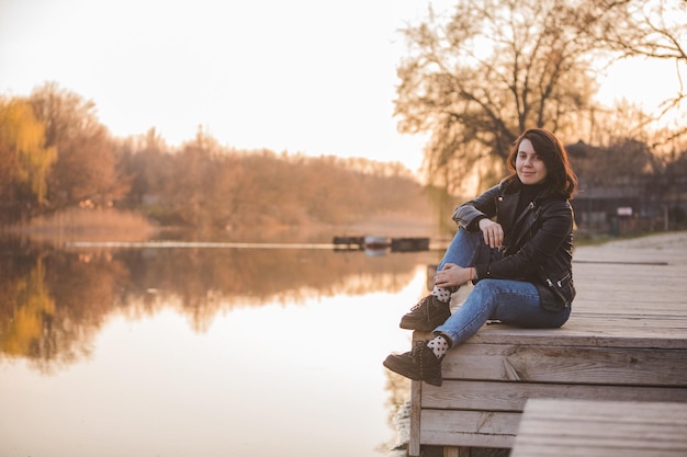 Mulher jovem e bonita moda sentada no cais no pôr do sol com bela vista do rio