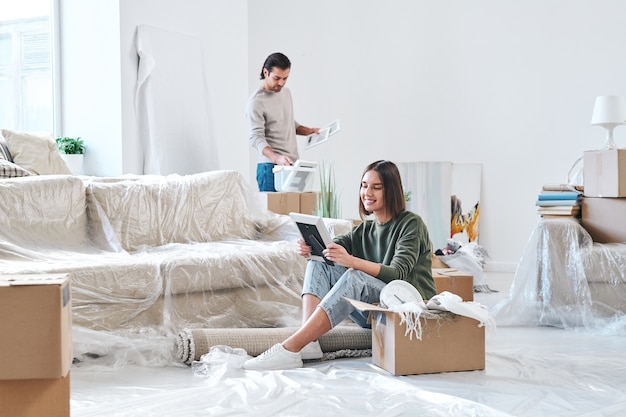 Mulher jovem e bonita morena olhando para a foto em moldura de madeira enquanto está sentada no chão de um apartamento ou casa nova