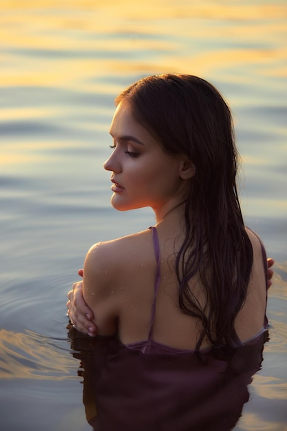 Mulher jovem e bonita na água do lago com vestido de verão ao pôr do sol Retrato de uma garota molhada romântica ao pôr do sol sol quente beleza natural de uma mulher