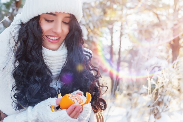 Foto mulher jovem e bonita na floresta de inverno descascando tangerina no dia ensolarado e gelado de natal ou ano novo