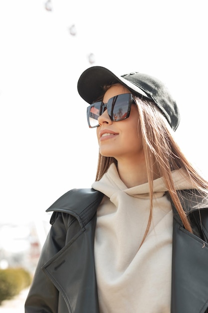 Mulher jovem e bonita na moda com óculos de sol elegantes legais em um capuz de jaqueta de couro preto de moda e boné preto moderno caminha em um dia ensolarado de primavera quente