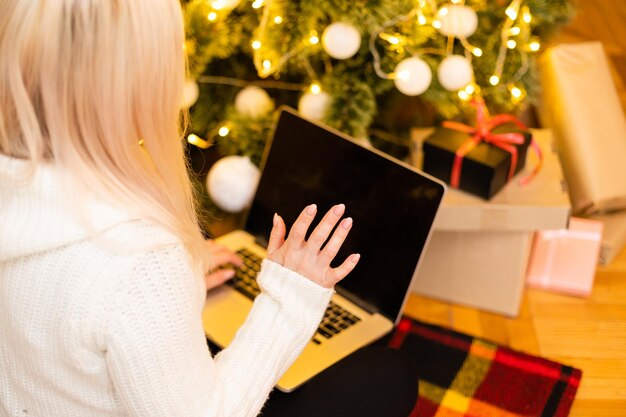 Mulher jovem e bonita na moda de inverno, sentada no chão com as pernas cruzadas, segurando o gadget de laptop na frente da decoração da árvore de Natal, enquanto olha para a câmera.