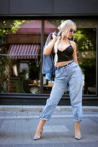 Foto mulher jovem e bonita na moda em óculos de sol, jeans e jaqueta posando na rua