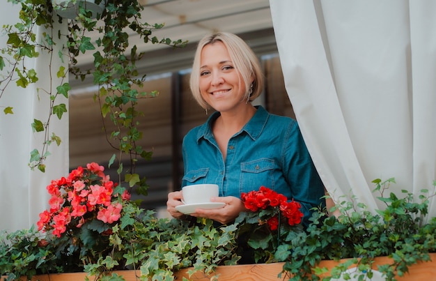 Mulher jovem e bonita na varanda com flores e cortina branca, desfrutando de uma xícara de café da manhã.