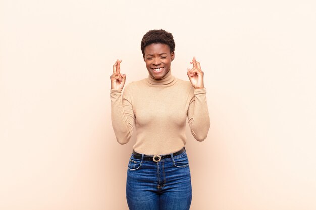 Mulher jovem e bonita negra, sentindo-se nervosa e esperançosa, cruzando os dedos, rezando e esperando a boa sorte