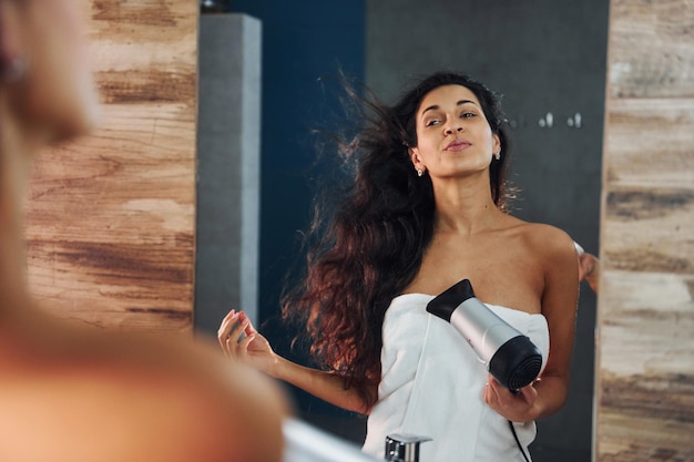 Foto mulher jovem e bonita no banheiro perto do espelho e secando o cabelo.