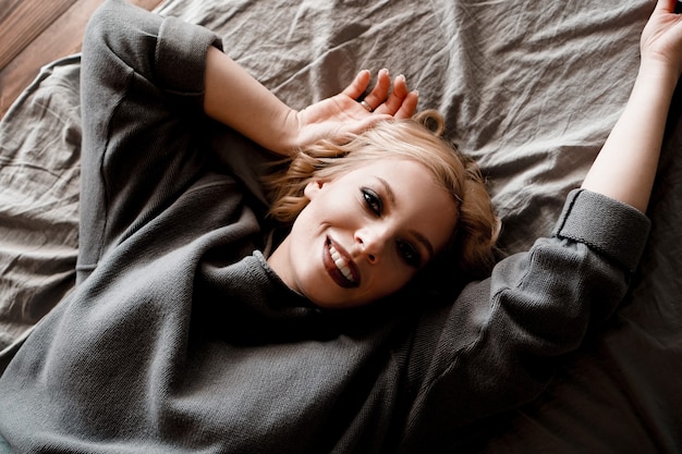 Mulher jovem e bonita no suéter cinza rindo deitada na cama em seu quarto aconchegante. Linda garota relaxando em casa. Manhã feliz