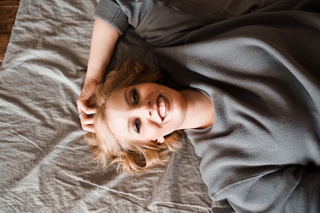 Mulher jovem e bonita no suéter cinza rindo deitada na cama em seu quarto aconchegante. linda garota relaxando em casa. manhã feliz