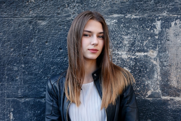 Mulher jovem e bonita no vestido e jaqueta de couro em pé sobre uma parede de pedra preta.