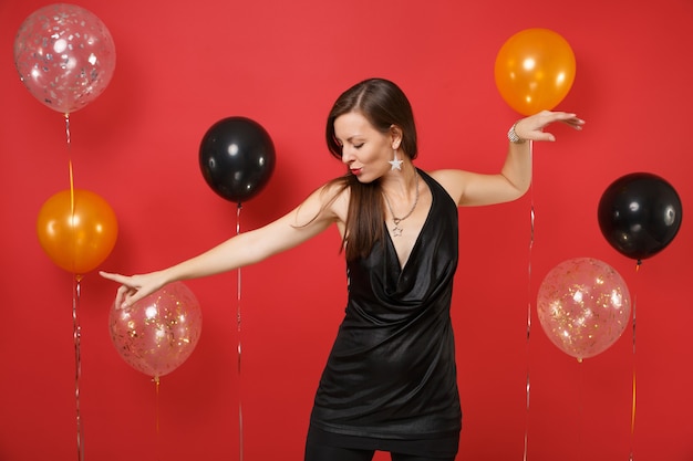Mulher jovem e bonita no vestido preto comemorando a dança espalhando as mãos, apontando o dedo em balões de ar de fundo vermelho brilhante. Dia das mulheres feliz ano novo aniversário maquete feriado festa conceito.