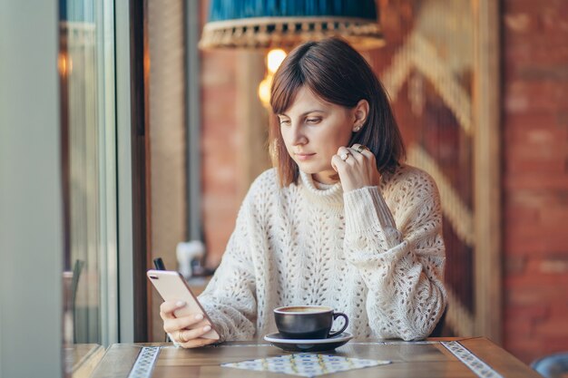 Mulher jovem e bonita numa camisola quente usando smartphone no café perto da janela com café. Aconchegante manhã de outono ou inverno.