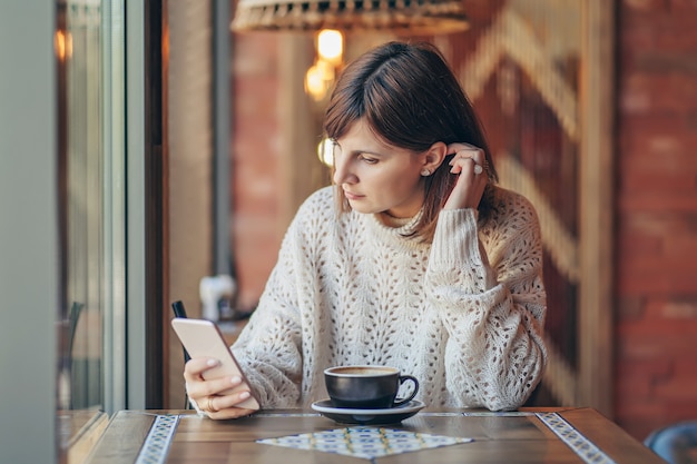 Mulher jovem e bonita numa camisola quente usando smartphone no café perto da janela com café. Aconchegante manhã de outono ou inverno.