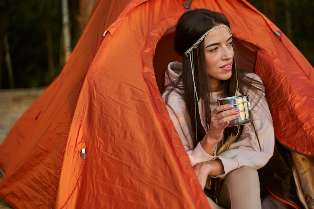 Mulher jovem e bonita olhando para longe e sorrindo enquanto segura uma caneca de bebida quente e está sentada dentro de uma barraca de acampamento laranja