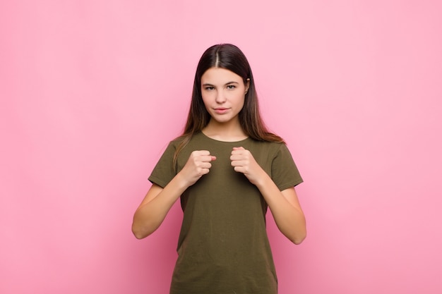 Mulher jovem e bonita parecendo confiante, irritada, forte e agressiva, com punhos prontos para lutar em posição de boxe contra a parede rosa