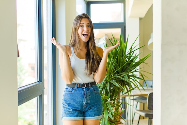 Foto mulher jovem e bonita parecendo feliz e animada, chocada com uma surpresa inesperada com as duas mãos abertas ao lado do rosto