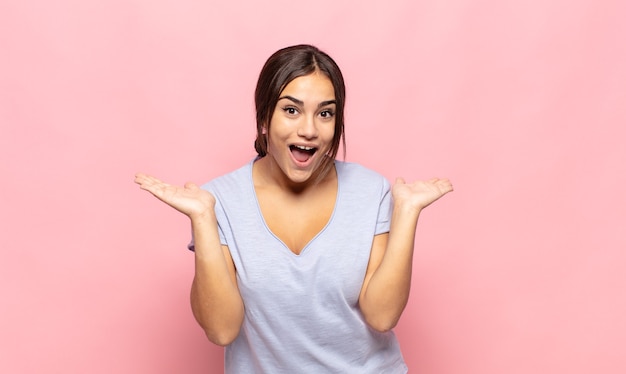 Mulher jovem e bonita parecendo feliz e animada, chocada com uma surpresa inesperada com as duas mãos abertas ao lado do rosto