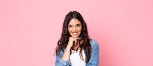 Mulher jovem e bonita parecendo feliz e sorrindo com a mão no queixo, pensando ou fazendo uma pergunta, comparando opções