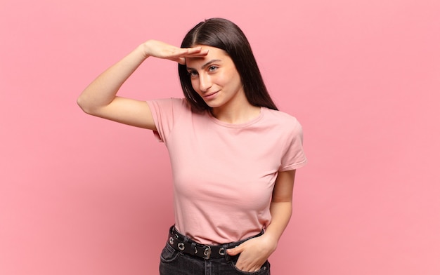 Mulher jovem e bonita parecendo perplexa e atônita, com a mão na testa olhando para longe, observando ou procurando