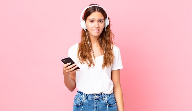 Mulher jovem e bonita parecendo perplexa e confusa com fones de ouvido e um smartphone