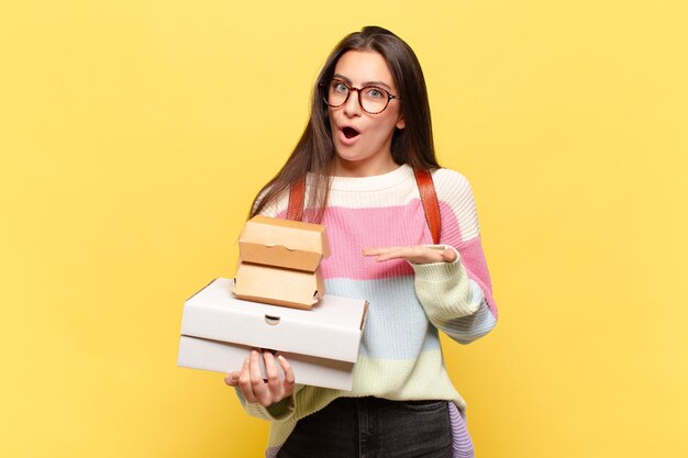 Mulher jovem e bonita parecendo surpresa e chocada, com o queixo caído segurando um objeto com a mão aberta na lateral. pegue um conceito de fast food fácil