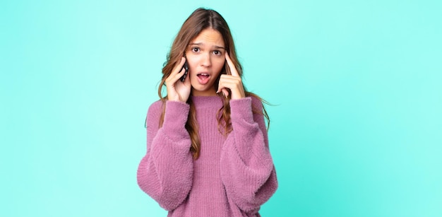 Mulher jovem e bonita parecendo surpresa, percebendo um novo pensamento, ideia ou conceito e usando um smartphone